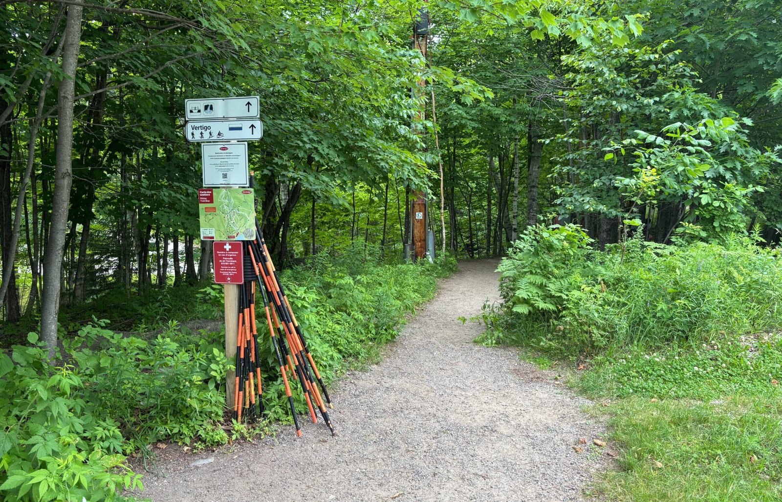Forest trail entrance with hiking signs, trail map, and orange-handled walking sticks at Mont-Tremblant’s Les Ruisseaux Loop.