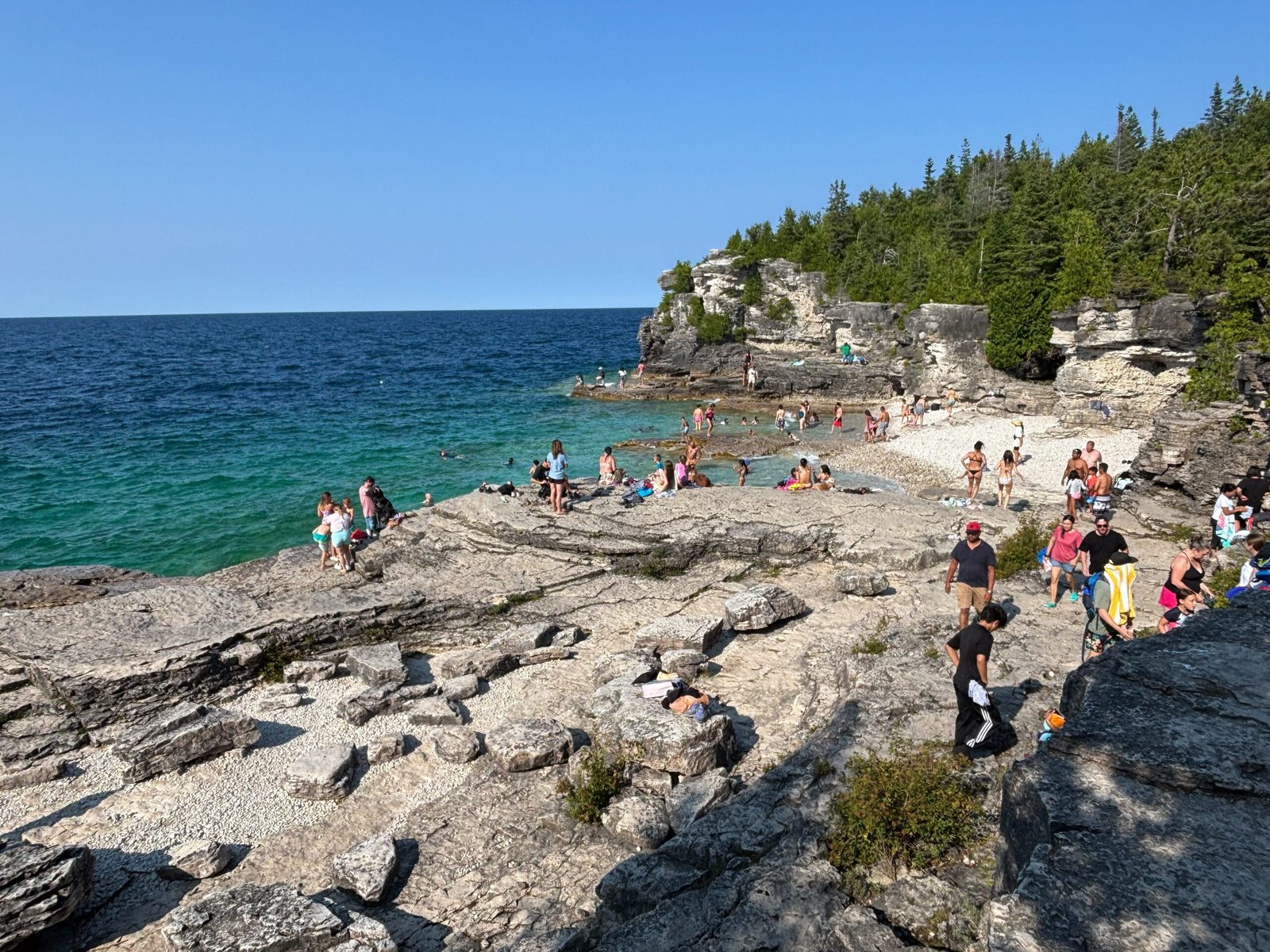Tourists swimming and relaxing along the rocky shore at the Grotto, Bruce Peninsula National Park, with clear turquoise water in the background.