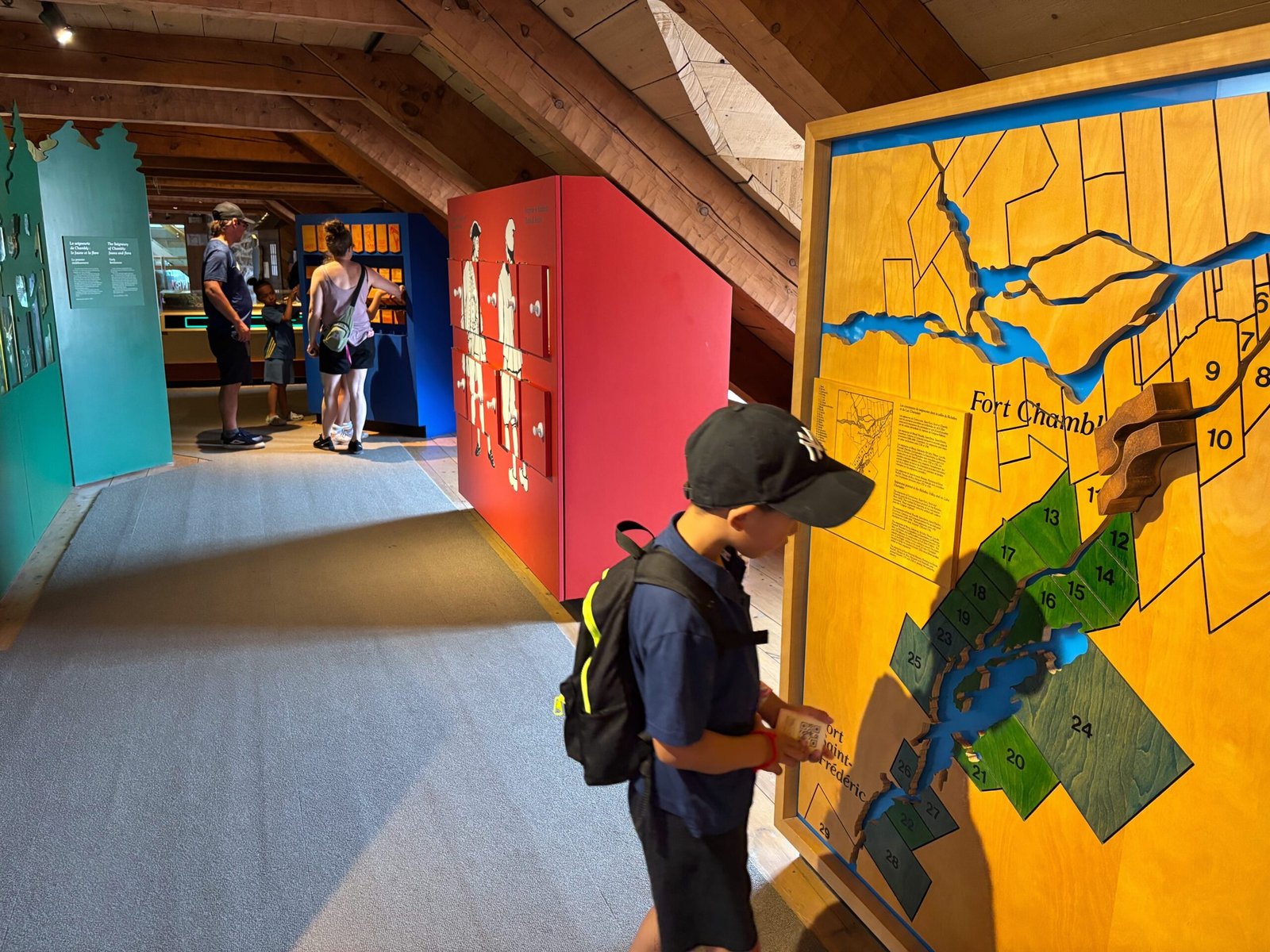 Child exploring interactive wooden map of Fort Chambly while families visit museum exhibits.