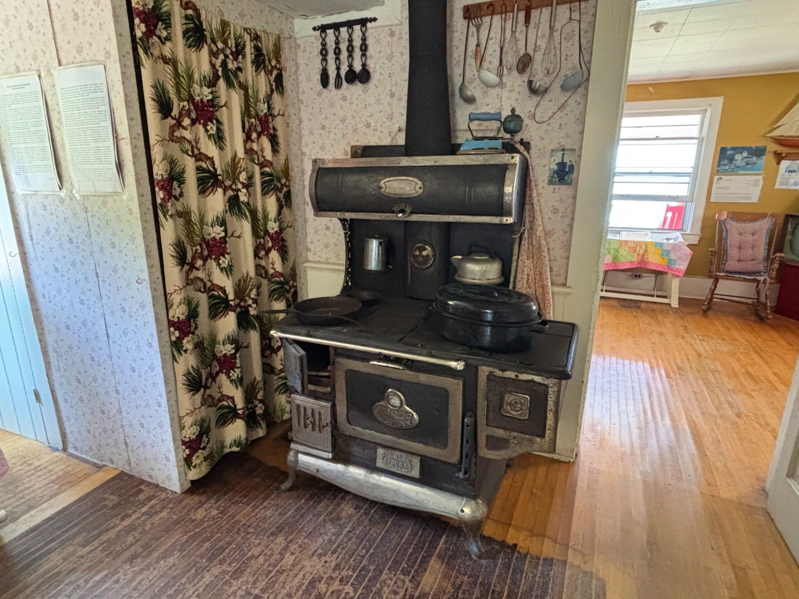 Antique cast-iron stove with pots and kettles, set in a kitchen with floral wallpaper.