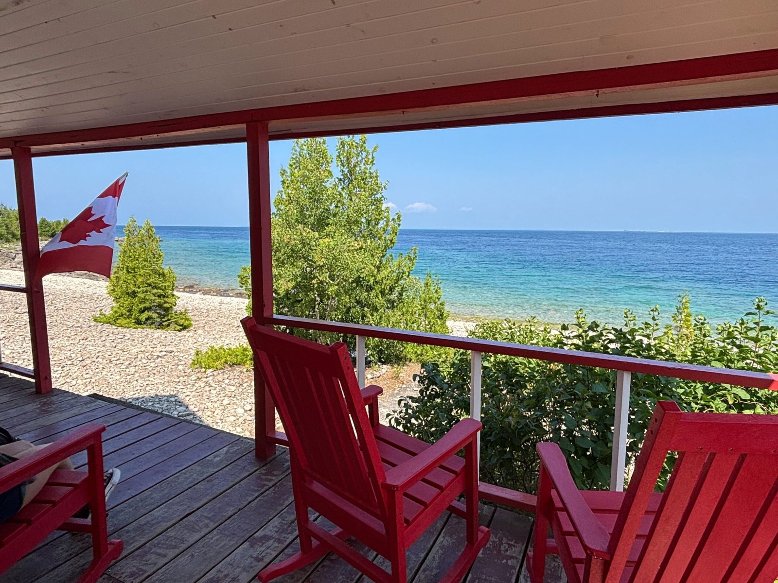 View from a red rocking chair on the Lightstation porch, showing a Canadian flag, pebble beach, trees, and turquoise water.