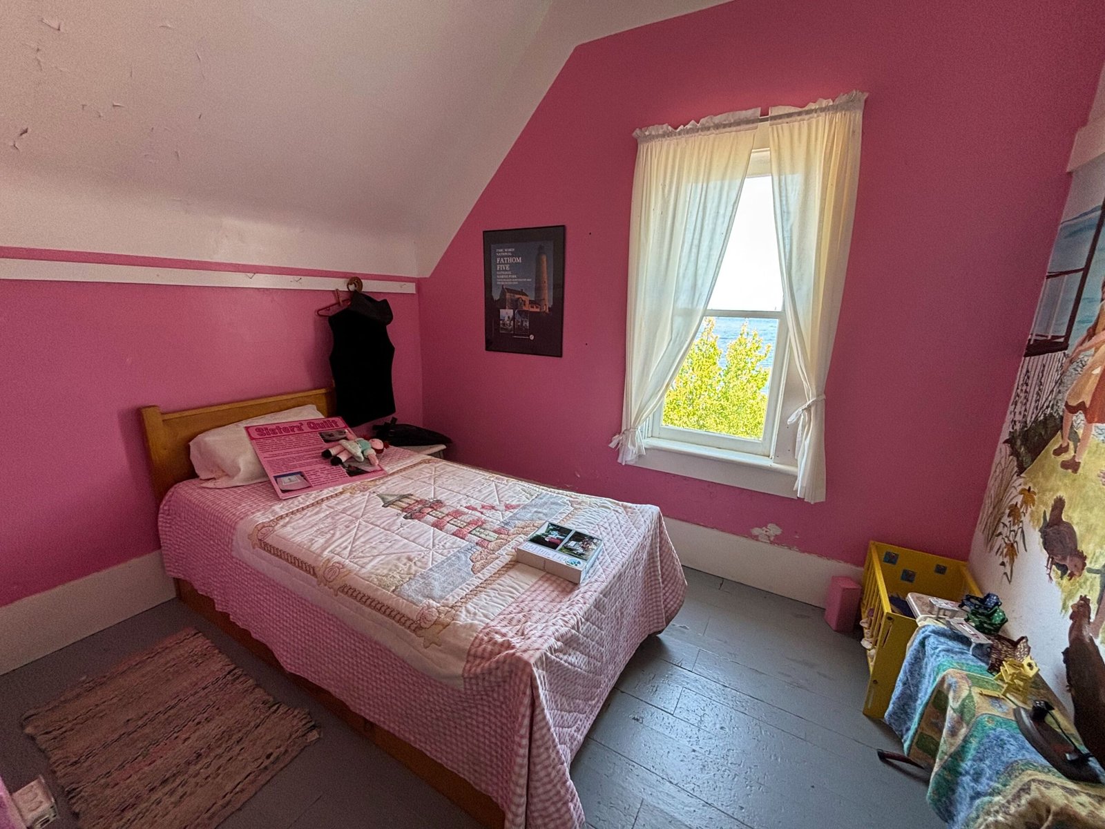Bedroom with pink walls and a single bed, decorated with a quilt and dolls, with a window view of the water.